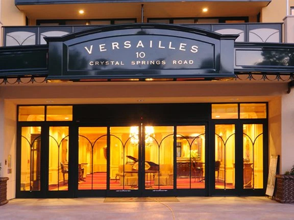 The lit glass entrance of the Versailles senior community at 10 Crystal Springs Road